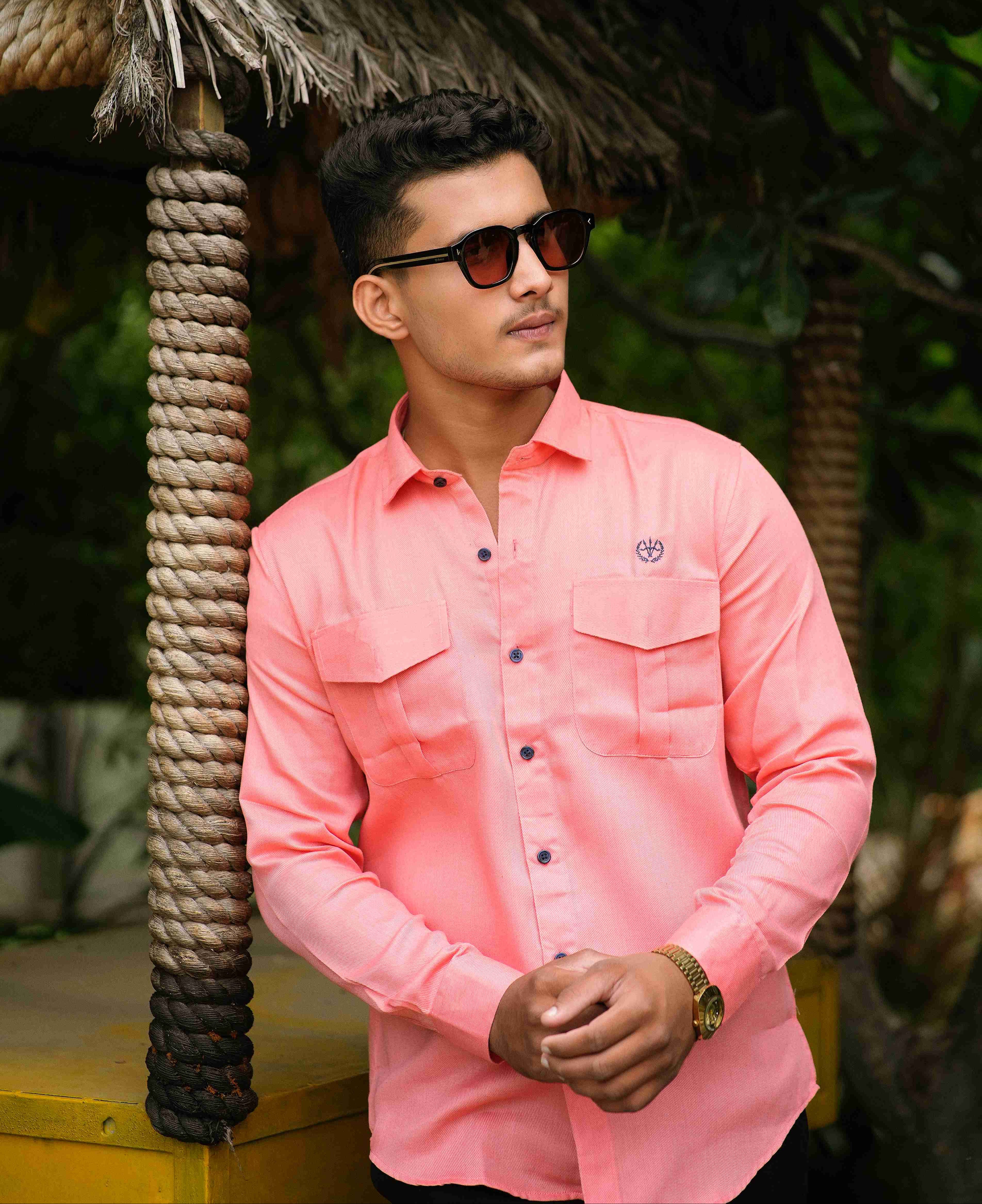 Man wearing a pink shirt and sunglasses standing under a thatched roof with greenery in the background