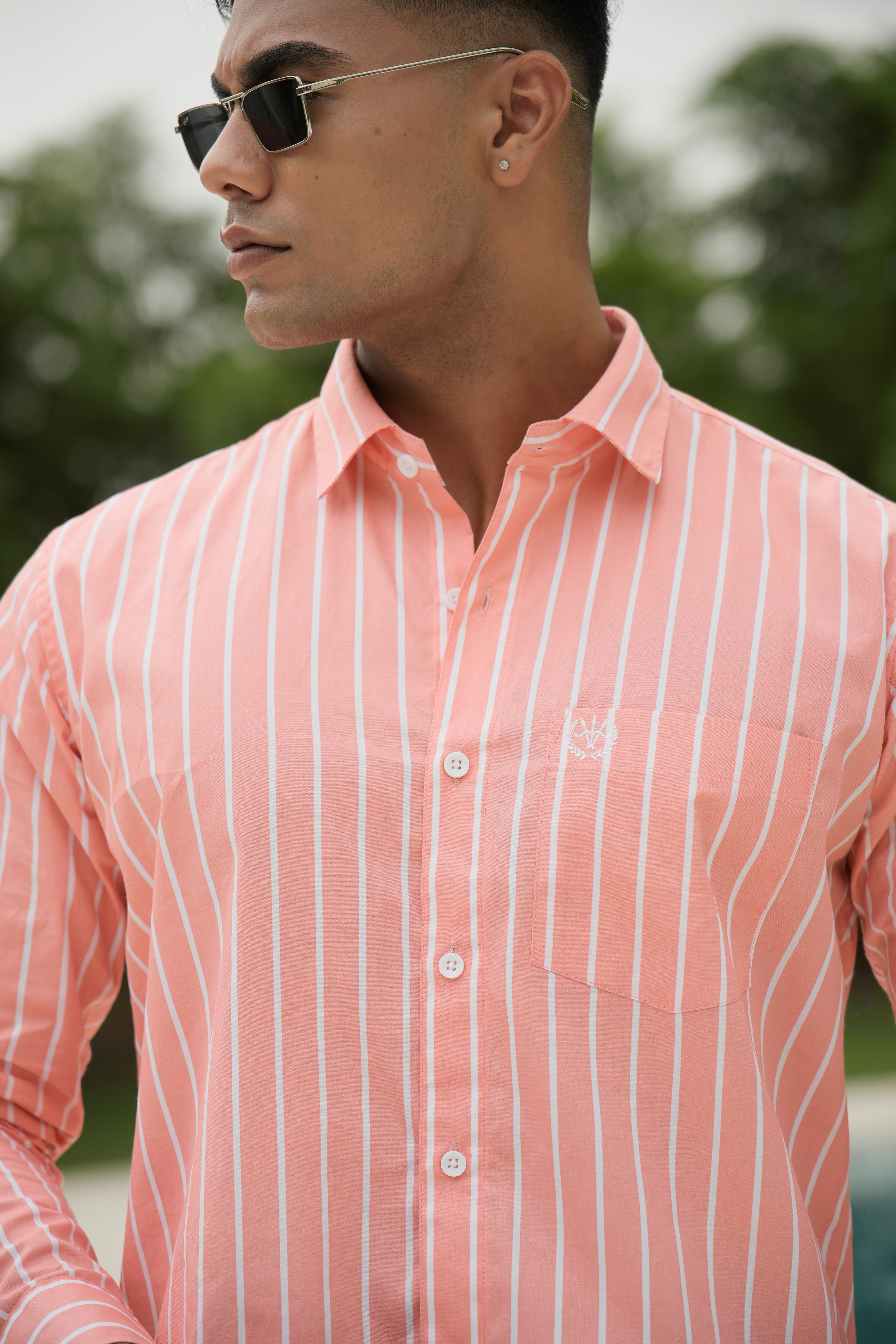 Amerald_arrows Man wearing a pink striped shirt with sunglasses, standing outdoors with blurred greenery in the background