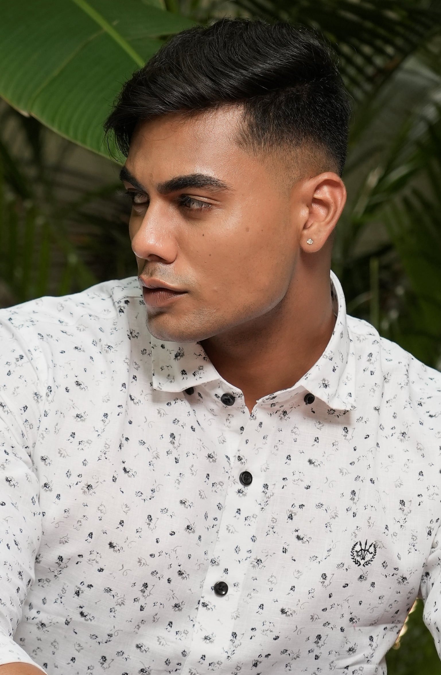 Person wearing a white patterned shirt and blue jeans standing indoors with plants in the background