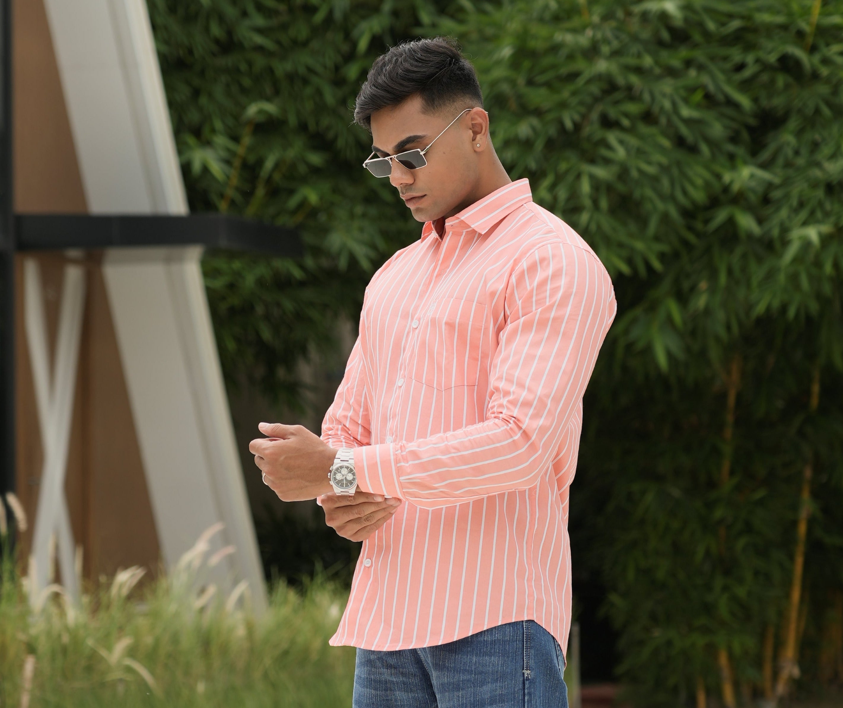 Amerald_arrows Person wearing a pink formal strip shirt  and blue jeans standing outdoors with trees and a building in the background.